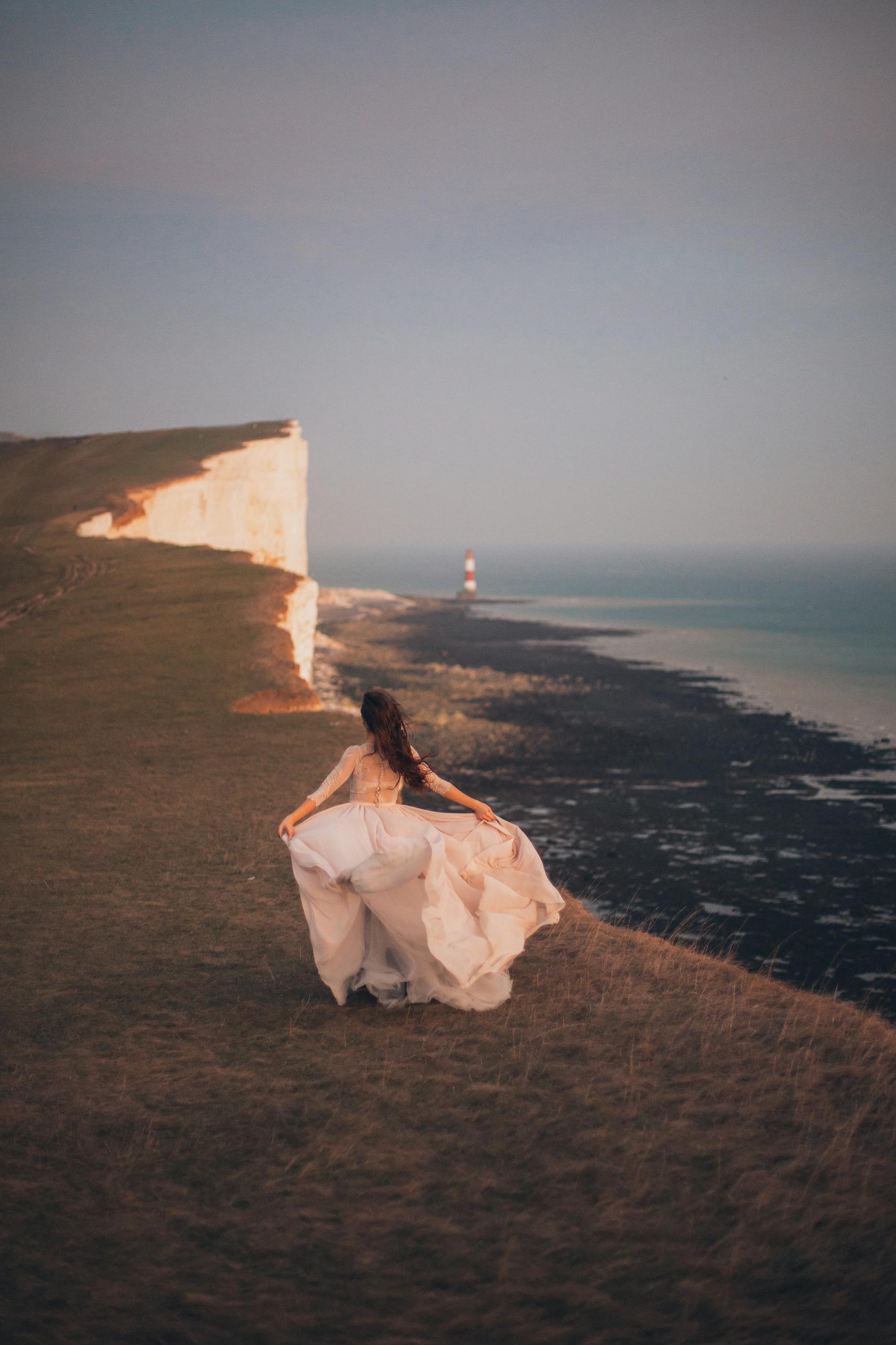 Bride in flowing dress on a dramatic cliff overlooking the lighthouse and sea.